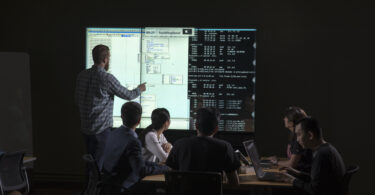 People gathered around table and man pointing to tech display